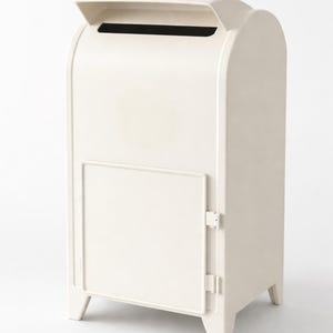 Op de afbeelding: Een cr&egrave;mekleurige, vrijstaande brievenbus met een gebogen bovenkant en een scharnierende deur. De brievenbus heeft een gleuf voor post aan de bovenkant en wordt ondersteund door vier korte poten. Het ontwerp is eenvoudig en modern.
