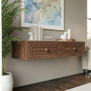 Puede incluir: Un mueble flotante de madera montado en la pared con dos cajones y pomos redondos. El mueble es de color marrón cálido y está montado en una pared de color neutro. En la parte superior se exhiben objetos decorativos, como libros y una figura de caballo de cerámica blanca.