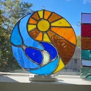 May include: A round stained glass suncatcher with a yin-yang design. The design features a yellow sun, blue water, and orange and brown accents. The suncatcher is displayed on a white windowsill with a blue sky background.