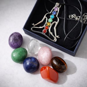 Könnte beinhalten: Eine silberne Anhängerkette mit einer sitzenden Figur mit sieben farbigen Edelsteinen, die Chakren darstellen, präsentiert in einer blauen Schachtel. Sieben polierte Edelsteine in verschiedenen Farben, darunter Lila, Grün und Rot, umgeben die Schachtel.