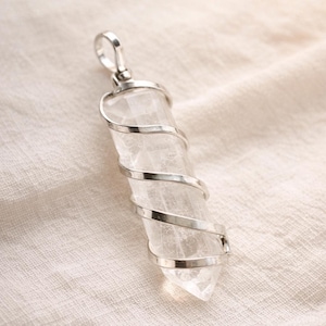 Può includere: Un ciondolo di cristallo di quarzo trasparente con un avvolgimento a spirale in metallo color argento e un anello nella parte superiore. Il cristallo è sfaccettato e ha un'estremità appuntita. Il ciondolo è esposto su un tessuto color crema.