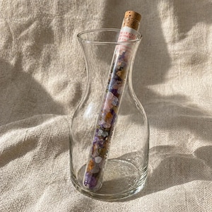 Könnte beinhalten: Eine klare Glasvase mit schmalem Hals enthält ein Glasröhrchen, das mit bunten Edelsteinen gefüllt ist. Das Röhrchen ist mit einem Korken verschlossen. Die Edelsteine sind eine Mischung aus lila, rosa, orangefarbenen und klaren Steinen.