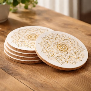 Op de afbeelding: Een stapel ronde onderzetters met een wit oppervlak en een gouden mandala-ontwerp. De onderzetters hebben een kurken basis en worden gepresenteerd op een houten oppervlak. De onderzetters zijn een decoratief woonaccessoire.