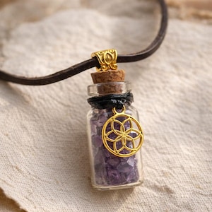Könnte beinhalten: Eine kleine Halskette mit einer Glasflasche, gefüllt mit lila Kristallen. Die Flasche hat einen Korken und einen goldfarbenen Anhänger mit floralem Design. Die Halskette hat ein braunes Lederband.