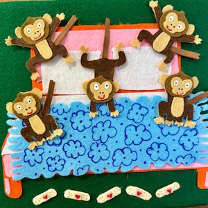 May include: A felt craft depicting five brown monkeys on a bed with a blue floral blanket. The bed has orange legs and a pink headboard. Band-aids with red hearts are at the bottom. The background is green.