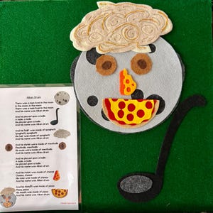 May include: A felt craft depicting a face with food features, including a pizza mouth, cheese nose, and spaghetti hair. The face is on a green background, with a black ladle and a printed poem titled "Aiken Drum".