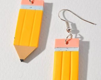 May include: A pair of novelty earrings shaped like yellow pencils. Each earring has a silver fish hook, a pink eraser, a light blue band, and a brown pencil tip with a black point. The earrings are a fun accessory.
