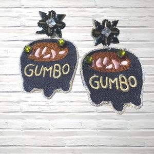 May include: A pair of handmade earrings featuring a dark blue, embroidered gumbo pot design. The earrings have a gold trim and the word "GUMBO" in gold thread. The top of the earrings have a black floral design with a clear crystal center.