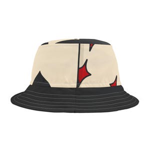 May include: A bucket hat with a black brim and a cream-colored crown. The crown features a repeating pattern of black shapes and red starburst designs. The hat is a casual accessory.