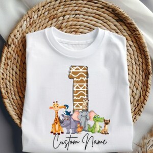 Könnte beinhalten: Weißes T-Shirt mit einer großen Zahl 1 im Giraffenmuster. Unter der Zahl befinden sich Zeichentricktiere wie Giraffe, Nashorn, Elefant, Krokodil und Erdmännchen. Der Text "Custom Name" ist kursiv geschrieben.