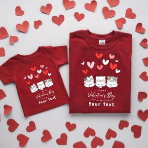 Könnte beinhalten: Zwei rote T-Shirts mit Valentinsthema. Jedes Shirt zeigt eine Grafik von drei Cartoon-Katzen mit herzförmigen Ballons. Der Text "Happy Valentine's Day" und "Your Text" ist darunter gedruckt. Rote Herz-Ausschnitte umgeben die Shirts.