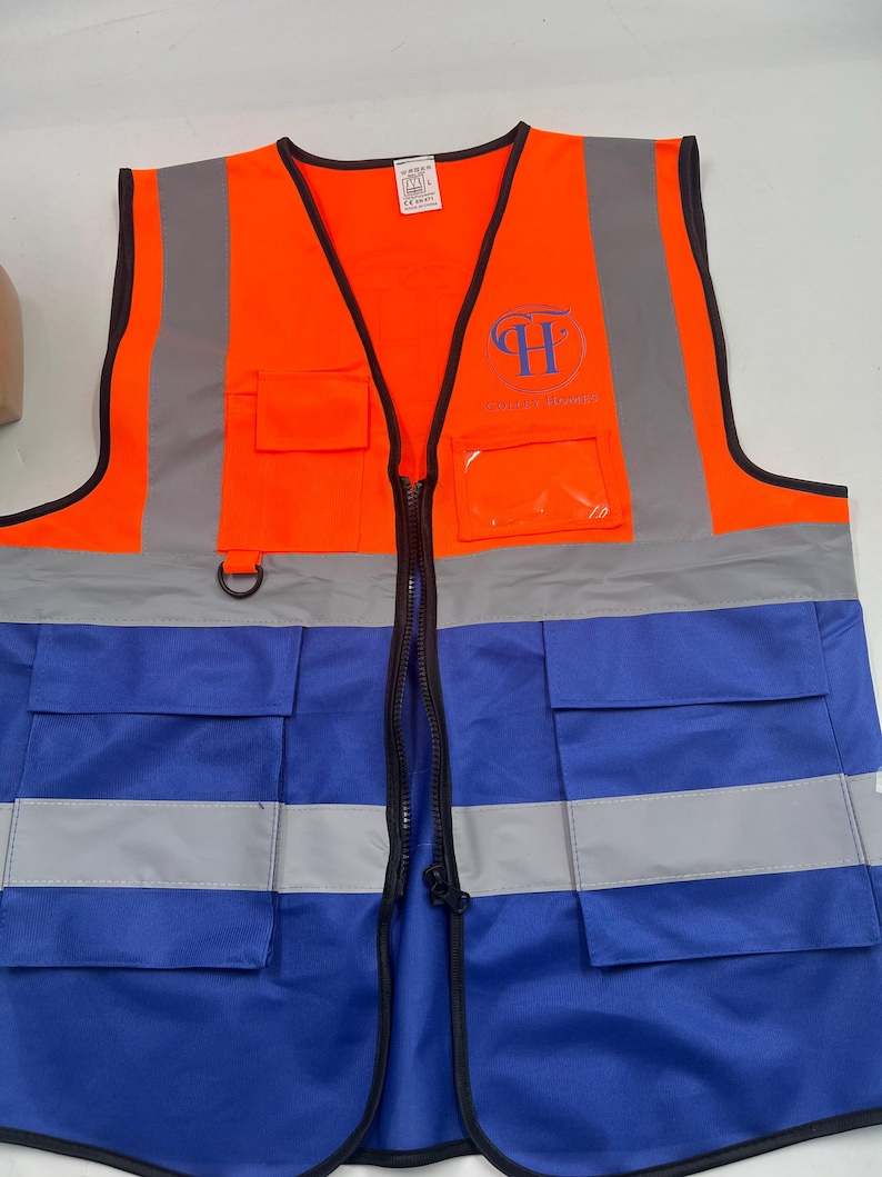 May include: High-visibility safety vest in bright orange and blue. The vest features reflective silver stripes and a black zipper. It has multiple pockets and a logo on the chest. The vest is designed for enhanced visibility.
