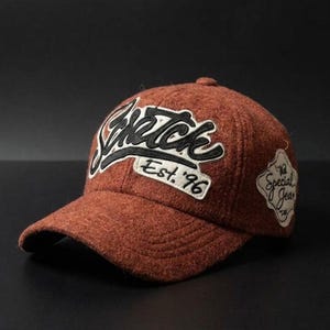 Puede incluir: Gorra de béisbol marrón con la palabra "Sketch" en escritura blanca y contorno negro. El texto "Est. '96" está debajo de la palabra "Sketch". Un pequeño parche en el lateral dice "Special Jean". La gorra tiene una visera curva.