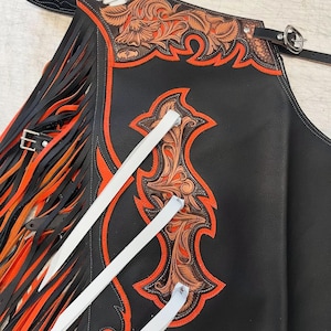 May include: Black leather chaps with orange and brown floral tooling and fringe. The chaps have white leather straps and a decorative arrow design. The edges are trimmed with orange stitching.