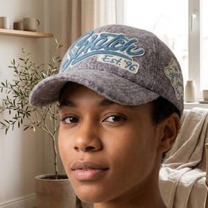 Könnte beinhalten: Eine graue, texturierte Baseballkappe mit einem großen, blau gestickten Schriftzug auf der Vorderseite mit dem Text Est. 96. Ein kleinerer dekorativer Aufnäher mit dem Text The Special Jean ist an der Seite der Kappe mit gebogenem Schirm zu sehen.