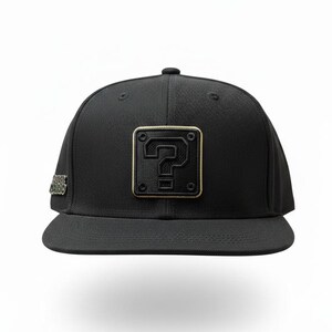 May include: A black baseball cap featuring a square patch on the front with a raised question mark icon outlined in gold. The side of the cap displays a metallic silver Super Mario logo emblem. The hat has a flat brim and a structured crown.