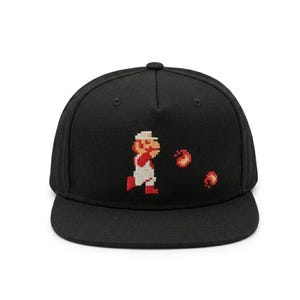 May include: A black baseball cap featuring an embroidered pixel art character wearing a white hat and red overalls, throwing two small orange and red fireballs. The cap has a structured crown, a flat brim, and a central button on the top.