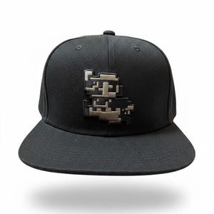 Pode incluir: Um boné de beisebol preto com um design bordado de personagem pixelizado em tons de castanho e bege no painel frontal. O boné possui uma copa estruturada, uma aba reta e um botão no topo, exibindo uma construção clássica de seis painéis.