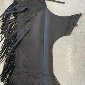 May include: A black leather chaps garment featuring long fringe along the side, an embossed cross design on the upper thigh, and intricate floral tooling at the top. The piece includes adjustable buckle straps at the waist and side for secure fastening.
