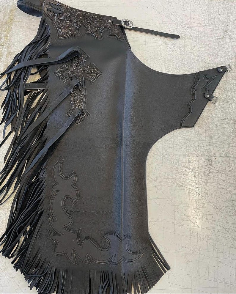 May include: A black leather chaps garment featuring long fringe along the side, an embossed cross design on the upper thigh, and intricate floral tooling at the top. The piece includes adjustable buckle straps at the waist and side for secure fastening.