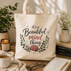 Puede incluir: Un bolso de tela color crema con un gráfico central que incluye el texto "it's a Beautiful mind thing" en letra cursiva negra y roja. El diseño presenta una ilustración detallada de un cerebro humano, hojas verdes y flores blancas.