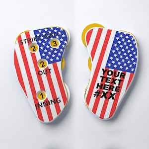 May include: Two baseball scoreboard clickers featuring a red, white, and blue American flag pattern. The left clicker displays STRIKE, BALL, OUT, and INNING with numerical dials. The right clicker features the text YOUR TEXT HERE #XX.