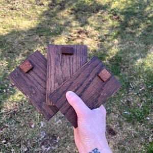 May include: Three dark brown wooden coasters with a square wooden accent piece attached. The coasters are held in a hand against a background of green grass.