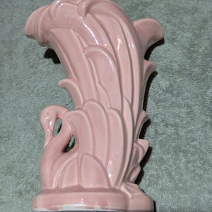 May include: A pink ceramic vase featuring a swan motif. The vase has a curved, stylized form with a swan on the front and a fan-like design at the back. The vase is a pale pink colour.