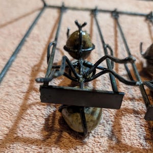 May include: A metal sculpture of an ant on a swing. The ant's body is a rounded stone, and the swing is made of black metal. The sculpture is set against a peach-colored background.
