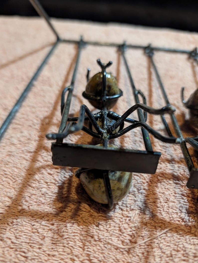 May include: A metal sculpture of an ant on a swing. The ant's body is a rounded stone, and the swing is made of black metal. The sculpture is set against a peach-colored background.