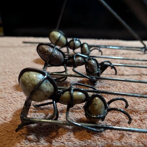 May include: Close-up of several metal ant sculptures. Each ant has a body made of a light-colored stone, encased in a dark metal wire frame. The ants have six legs and are arranged in a row.