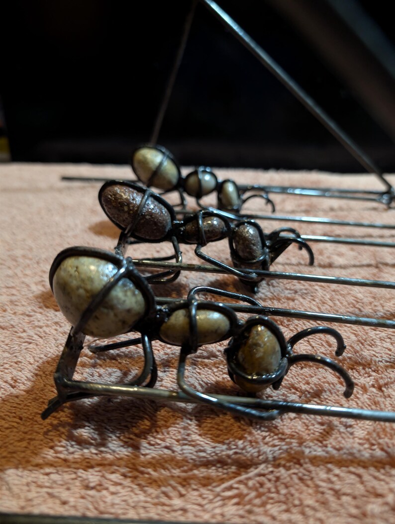 May include: Close-up of several metal ant sculptures. Each ant has a body made of a light-colored stone, encased in a dark metal wire frame. The ants have six legs and are arranged in a row.