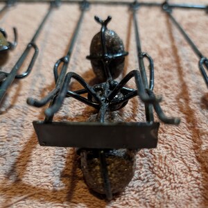 May include: Close-up of a metal sculpture of an ant. The ant is black with a textured body and legs. It is attached to a series of metal rods. The sculpture is on a light brown surface.