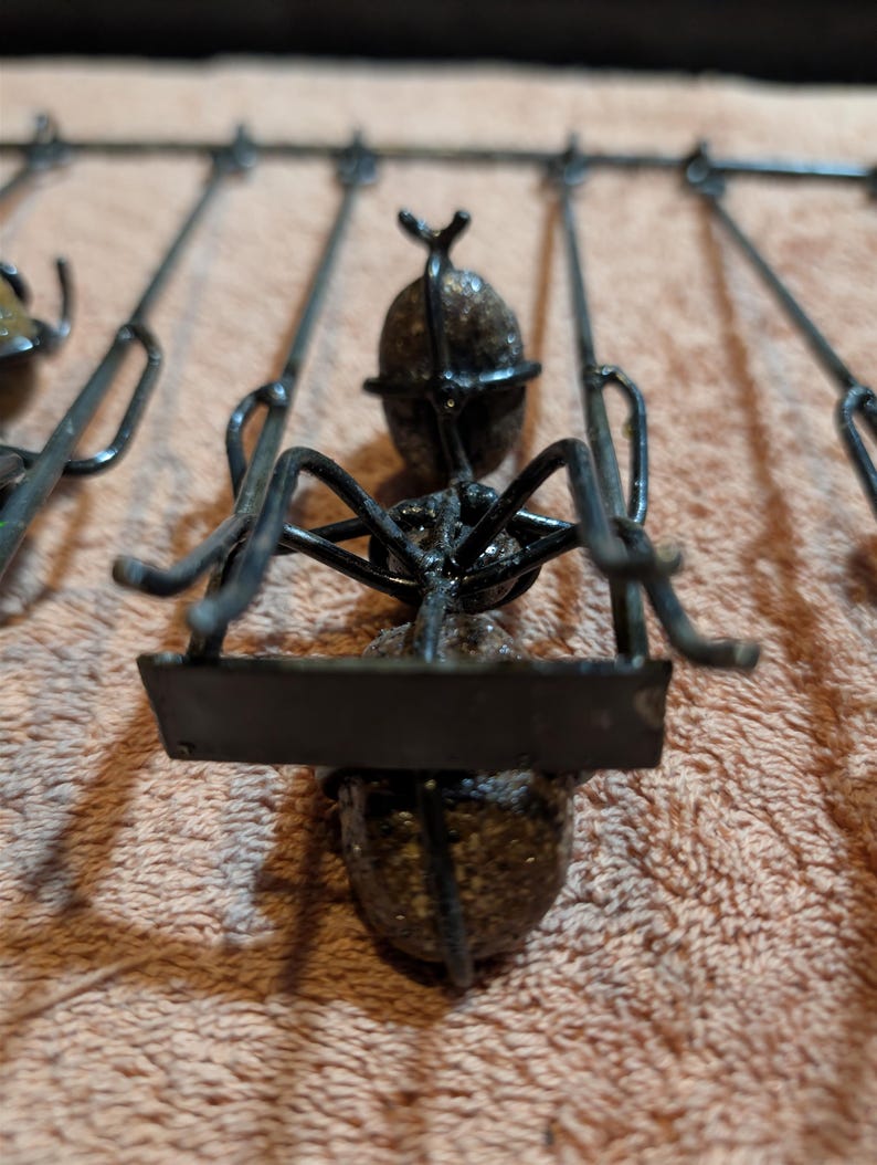 May include: Close-up of a metal sculpture of an ant. The ant is black with a textured body and legs. It is attached to a series of metal rods. The sculpture is on a light brown surface.