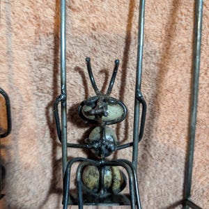 May include: A metal ant sculpture with a whimsical design. The ant's body is made of rounded stones, and its legs and antennae are crafted from bent metal rods. The sculpture is a unique piece of home decor.