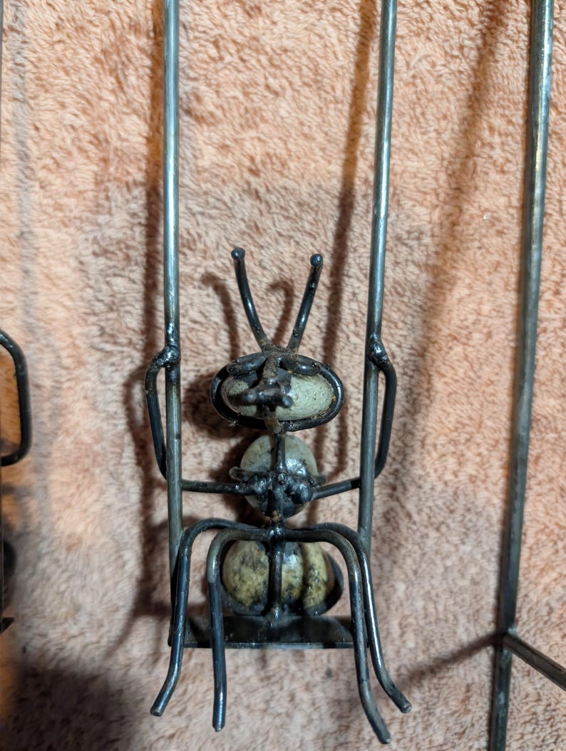May include: A metal ant sculpture with a whimsical design. The ant's body is made of rounded stones, and its legs and antennae are crafted from bent metal rods. The sculpture is a unique piece of home decor.