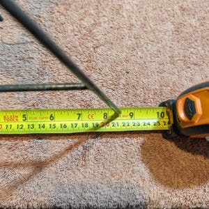 May include: A black metal object is measured with a yellow tape measure. The tape measure shows a measurement of approximately 8 inches. The tape measure is orange and black. The background is a brown, textured surface.
