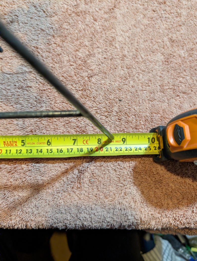 May include: A black metal object is measured with a yellow tape measure. The tape measure shows a measurement of approximately 8 inches. The tape measure is orange and black. The background is a brown, textured surface.