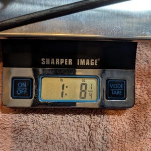 May include: A digital scale with the text "SHARPER IMAGE" displays a weight of 1 pound and 8 1/4 ounces. The scale has an on/off button and a mode/tare button. The background is a light pink towel.