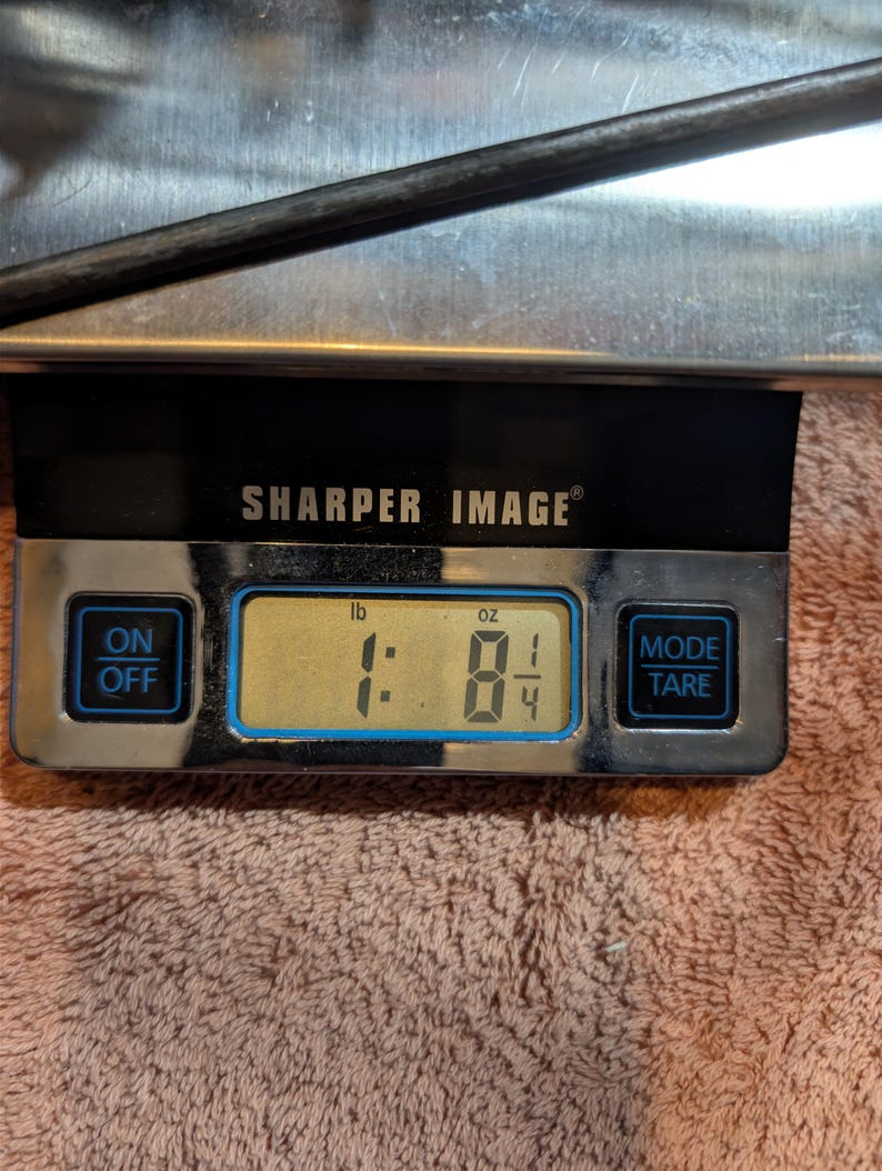 May include: A digital scale with the text "SHARPER IMAGE" displays a weight of 1 pound and 8 1/4 ounces. The scale has an on/off button and a mode/tare button. The background is a light pink towel.