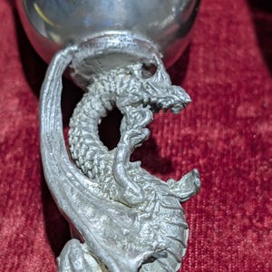 May include: A silver-colored goblet with a detailed dragon sculpted as the stem. The dragon's wings and body wrap around the cup, with the dragon's head near the rim. The goblet is set against a red background.