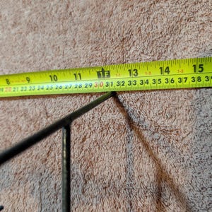May include: A close-up of a yellow measuring tape with black markings, showing measurements in inches. The tape is positioned near a dark metal object. The background is a textured, light brown surface.