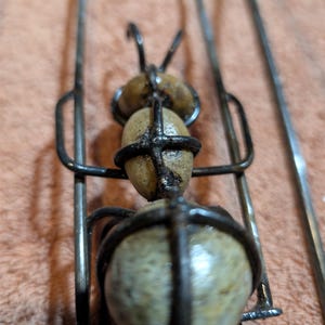 May include: A handcrafted metal ant sculpture with three stone-like spheres for the body and head. The ant's body is held together by a dark metal frame, with thin metal legs and antennae. The sculpture is on a neutral background.