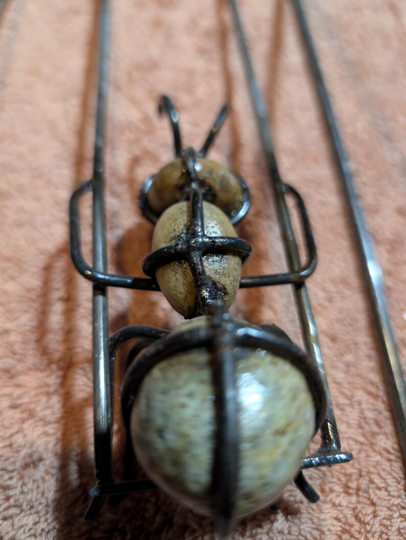 May include: A handcrafted metal ant sculpture with three stone-like spheres for the body and head. The ant's body is held together by a dark metal frame, with thin metal legs and antennae. The sculpture is on a neutral background.