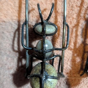 May include: A metal ant sculpture with three speckled stone-like spheres held within a wireframe body. The ant has curved antennae and long legs. The sculpture is set against a textured, light brown background.