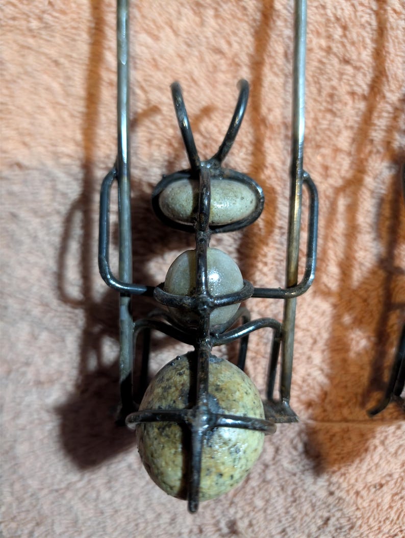 May include: A metal ant sculpture with three speckled stone-like spheres held within a wireframe body. The ant has curved antennae and long legs. The sculpture is set against a textured, light brown background.