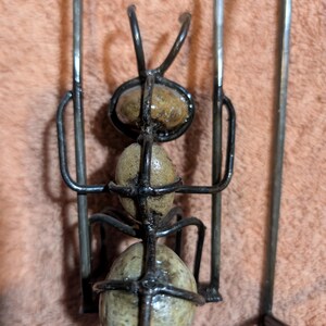 May include: A handcrafted metal ant sculpture with a wire frame and three stone-like spheres for the body. The ant has curved antennae and is set against a textured, peach-colored background.