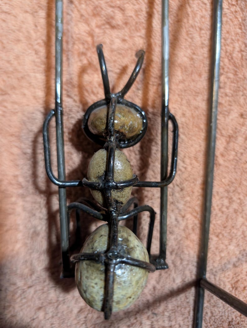 May include: A handcrafted metal ant sculpture with a wire frame and three stone-like spheres for the body. The ant has curved antennae and is set against a textured, peach-colored background.