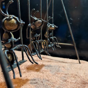 May include: A metal sculpture featuring three stylized ants suspended within a cage-like structure. The ants have detailed bodies and antennae, with a mix of bronze and dark metal finishes. The sculpture is set against a neutral background.