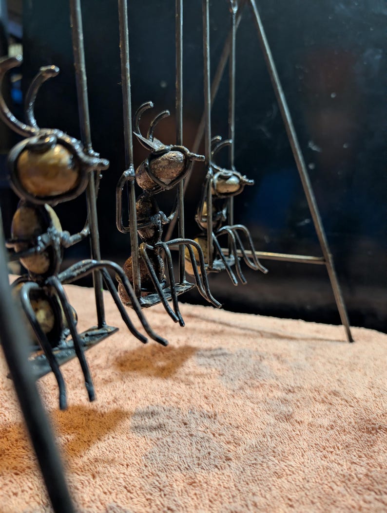 May include: A metal sculpture featuring three stylized ants suspended within a cage-like structure. The ants have detailed bodies and antennae, with a mix of bronze and dark metal finishes. The sculpture is set against a neutral background.